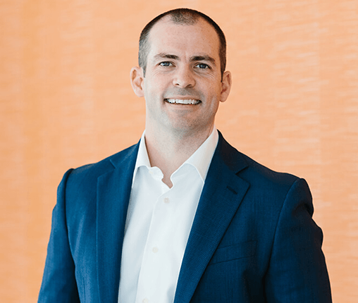 AboutUs-Leadership_3070_BrianFeichter A man in a blue suit and white shirt smiles in front of an orange background. His hair is short, and he stands with both arms relaxed at his sides.