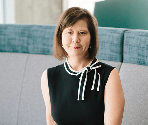 AboutUs-Leadership_3070_CharleneYork A woman with shoulder-length brown hair is wearing a sleeveless black dress with a white bow at the collar. She is sitting on a gray and teal bench, smiling slightly at the camera.