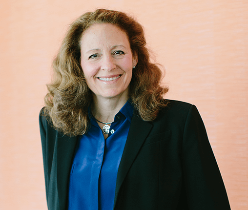 AboutUs-Leadership_3070_DollyWagnerWilkins Smiling woman with curly hair, wearing a blue blouse and black blazer, standing against a peach-colored background.