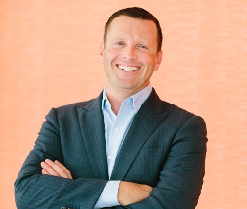AboutUs-Leadership_3070_Dustin_Wesley A man in a blue suit and light blue shirt is smiling with arms crossed against an orange background.