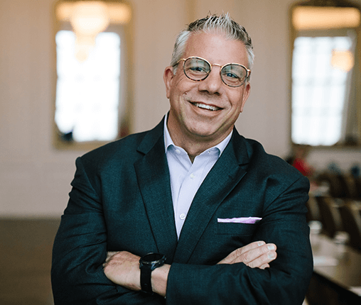 AboutUs-Leadership_3070_JoeGuerrisi A man with short gray hair, wearing glasses and a dark suit with a light dress shirt, smiles with arms crossed in a well-lit room.