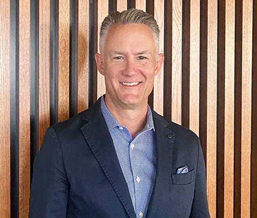 AboutUs-Leadership_3070_JoeKniple A person in a dark blazer and blue polka dot shirt smiles while standing in front of a wooden slat wall.