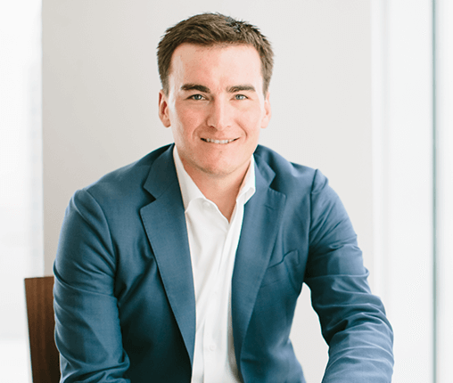 AboutUs-Leadership_3070_JoelClum A man in a blue suit and white shirt is sitting and smiling at the camera. He has short brown hair and is positioned against a light, neutral background.