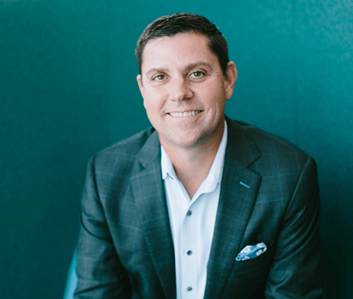 AboutUs-Leadership_3070_MikeGrayson A smiling man in a suit sits against a teal background, exuding confidence like the perfect arrangement of card modules in a sleek design. Hes donning a dark plaid jacket, crisp white shirt, and pocket square—a harmonious blend of style and elegance.