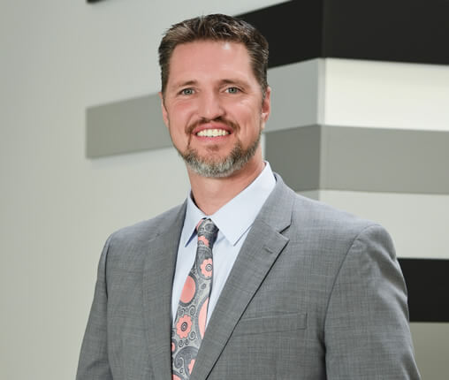 AboutUs-Leadership_3070_Mike_Sutter A man with short brown hair and a beard smiles at the camera. He is wearing a gray suit, light blue shirt, and a patterned tie. The background features a wall with alternating black, gray, and white stripes.