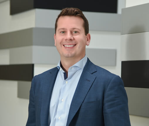 AboutUs-Leadership_3070_Ross_Spanier Smiling man in a blue suit and light blue shirt stands in front of a geometric patterned wall featuring shades of gray and black rectangles.