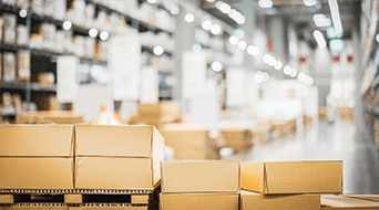 A warehouse interior with stacks of cardboard boxes on a wooden pallet in the foreground. The background features shelves filled with more boxes and soft, diffused lighting, creating a spacious and organized atmosphere.