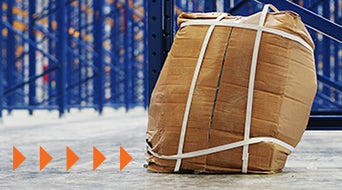 A crumpled cardboard box secured with white straps sits on a warehouse floor. Blue metal shelving is visible in the background. Orange arrows point to the left, indicating movement.
