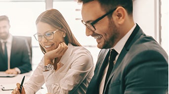 Two people in business attire are smiling and looking at a document or screen in an office setting. The woman has long hair and glasses, while the man has short hair and a beard. Another person is blurred in the background.
