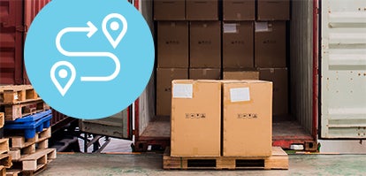 A warehouse scene with cardboard boxes stacked on a pallet in front of an open shipping container. A blue circle with a white route map icon is overlaid on the left side of the image. Wooden pallets are visible nearby.