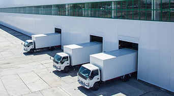 Three white delivery trucks are backed into loading docks at a large industrial warehouse. The building has a long row of windows above the docks, and the ground is paved with concrete.