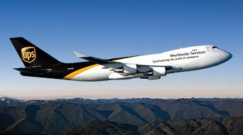 A UPS cargo airplane is flying over a mountainous landscape. The aircraft has a white body and black tail with the UPS logo, and the text Worldwide Services is visible on its side. The mountains below are covered with greenery.