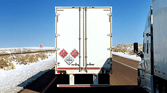 A white truck with hazard signs is on a snowy highway, traveling alongside a semi-truck on a clear day. The road curves slightly to the left, and the surroundings are covered in snow.