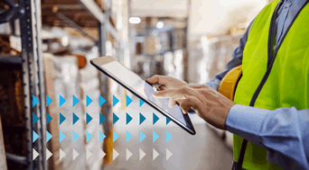 A person in a high-visibility vest uses a tablet to manage shipping invoices in a warehouse. The background shows shelves with boxes, while blue and gray arrow graphics overlay the image, suggesting movement or progress.