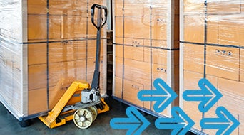 A pallet jack positioned to move large stacks of shrink-wrapped cardboard boxes in a storage area. Blue arrows are superimposed, pointing to the right, suggesting movement or direction.