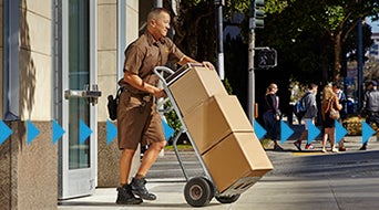 A delivery person in a brown uniform pushes a hand truck with cardboard boxes on a city sidewalk. The background shows pedestrians walking and a street intersection under a clear sky.