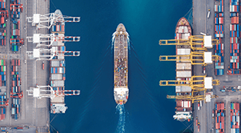 Aerial view of a cargo ship sailing through a narrow channel between two container terminals. The terminals are lined with cranes and stacks of colorful shipping containers on either side of the water.
