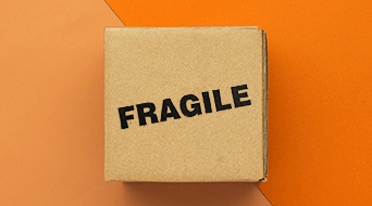 A small brown cardboard box with the word FRAGILE printed in bold black letters on top. The box is placed on a surface divided into two shades of orange, creating a contrasting background.