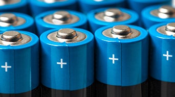 Close-up of several blue and silver batteries in a grid pattern. Each battery top displays a plus sign, indicating the positive terminal. The image focuses on the battery tops in sharp detail.