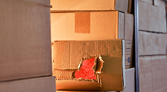Stacked cardboard boxes with one showing visible damage, revealing red packing material inside. The boxes are arranged closely together, suggesting a storage or warehouse setting.