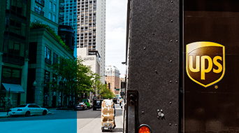 A UPS delivery truck is parked on an urban street. A hand truck loaded with packages is visible, while city buildings line the street. Several cars are parked nearby.