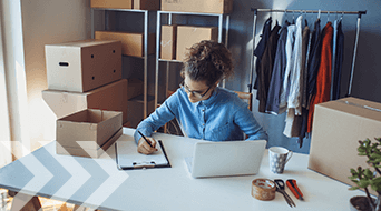 A person wearing glasses and a blue shirt writes on a clipboard at a desk with a laptop. Surrounding them are cardboard boxes and a clothing rack. Scissors and a coffee mug are on the desk.