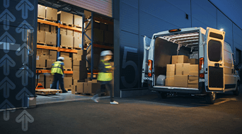 A delivery van is parked outside a warehouse with its back doors open. Workers wearing high-visibility vests and hard hats are moving boxes from the van into the warehouse. The area is dimly lit, with stacked cardboard boxes inside.
