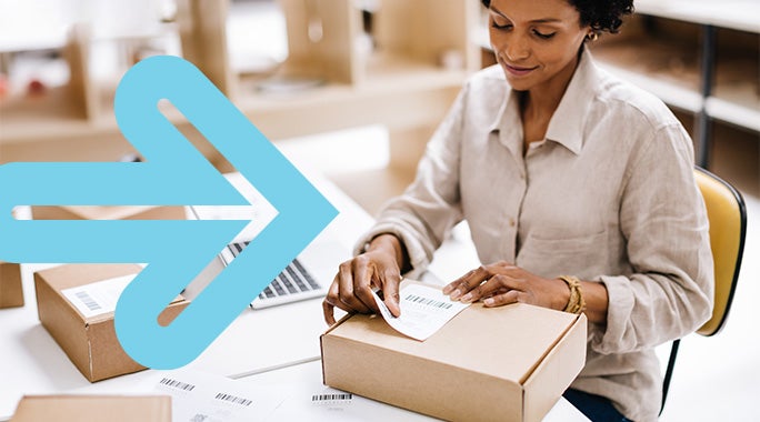 A woman sits at a desk, placing a barcode label on a cardboard box. She has short curly hair and wears a beige shirt. A large blue arrow overlays the image, pointing to the right. A laptop is partially visible in the background.