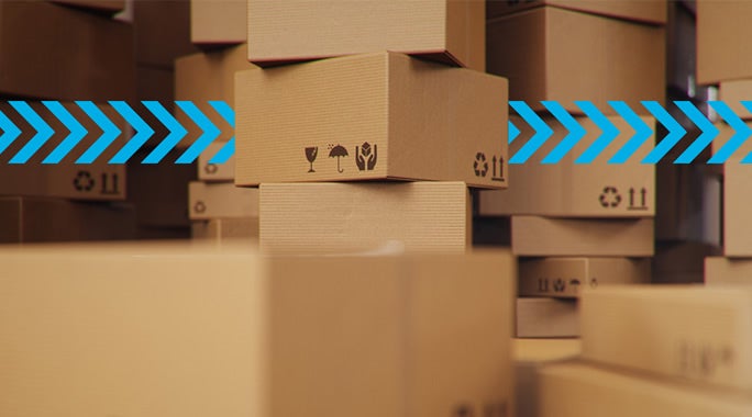 Stacks of brown cardboard boxes with various symbols, including fragile and recycling icons, in a warehouse setting. Blue arrows overlay the image, pointing to the right.