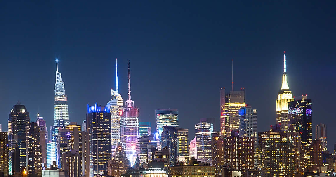 A nighttime view of a city skyline with brightly lit skyscrapers against a dark blue sky, including several tall buildings with illuminated spires and colorful lights.