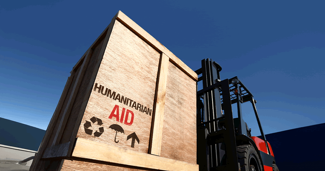 A large wooden crate labeled “Humanitarian Aid” is being lifted by a forklift against a clear blue sky. The crate features recycling, umbrella, and up arrow symbols.