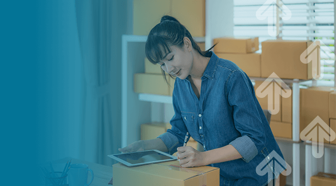 A woman in a denim shirt writes on a cardboard box while using a tablet, surrounded by other packages, suggesting she is processing online orders in a home office. White upward arrows are overlaid on the image.