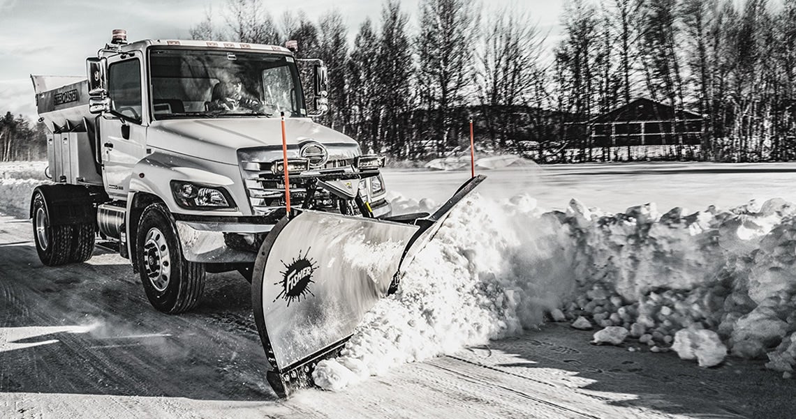 A snow plow truck clearing a snow-covered road, pushing snow to the side. Trees and a building are visible in the background on a cold, winter day.