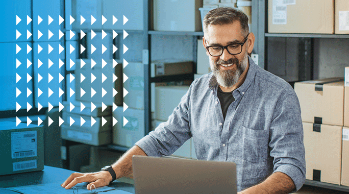 A man with a beard and glasses smiles while working on a laptop in a warehouse filled with cardboard boxes. White arrow graphics are overlaid on the left side of the image.