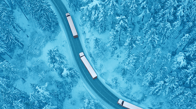 Aerial view of three trucks driving on a winding road through a snow-covered forest with trees densely packed on both sides.