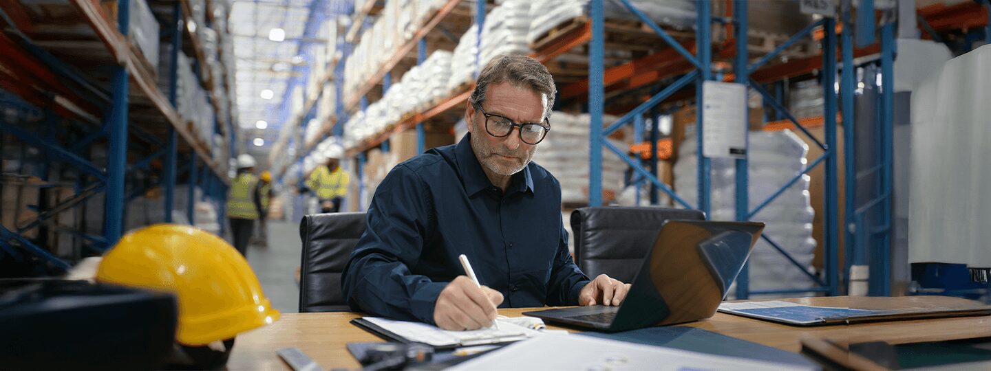 A man wearing glasses sits at a desk in a warehouse, writing in a notebook with his laptop open, possibly reviewing freight terms. Shelves stocked with goods and workers in safety vests are visible in the background.