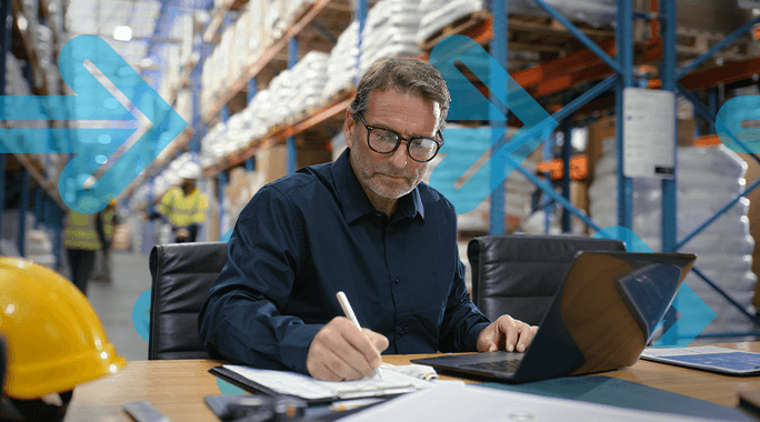 A man with glasses works at a desk in a warehouse, writing in a notebook with freight terms beside his open laptop. Shelves filled with bags are in the background, and blue arrows are overlaid on the image.