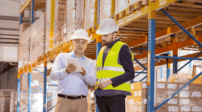 Two men wearing hard hats and safety vests stand in a warehouse, discussing a Blind Shipping Strategy displayed on a tablet. Shelves stocked with boxes are visible in the background.