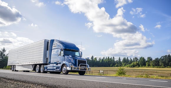 An ADA Enterprises blue semi-truck with a white trailer drives on a rural road bordered by green fields and trees under a partly cloudy sky, showcasing reliable B2B logistics.