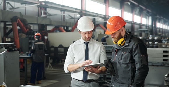 Two men from ADA Enterprises, specializing in B2B logistics, stand in a factory wearing safety helmets and work clothes, examining a tablet. One is in a tie and white helmet; the other wears a dark uniform with an orange helmet and yellow ear protectors.