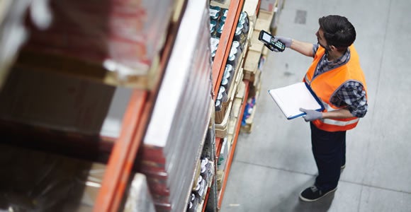 A warehouse worker in an orange safety vest scans items on a high shelf with a handheld device while holding a clipboard, managing CBD shipping for Green Roads in an aisle surrounded by stocked shelves.