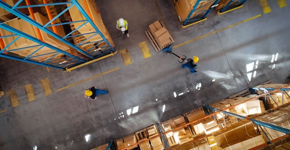Aerial view of a warehouse with shelves of boxes, three workers in safety vests and helmets moving around, and one handling Green Roads CBD shipping boxes with a pallet jack along the aisle.