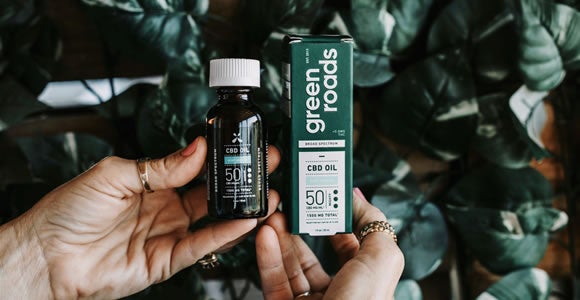 A person holds a bottle of CBD oil in one hand and a Green Roads CBD oil box in the other, surrounded by leafy green plants, highlighting trusted Green Roads products and convenient CBD shipping.