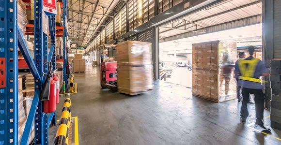 A forklift moves a pallet of boxes inside a bright warehouse, while two workers in safety vests stand nearby. Shelving and stacked pallets line the walls, streamlining peak season shipping as sunlight streams through upper windows.