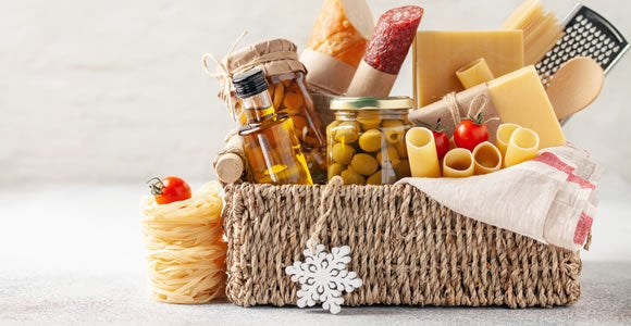 A woven basket filled with Italian food items—including olive oil, pasta, cheese, bread, salami, olives in jars, and cherry tomatoes—makes the perfect gift for peak season shipping. Uncooked pasta and a snowflake ornament sit in front of the basket.