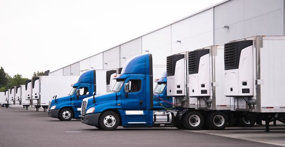 Several blue semi-trucks with white refrigerated trailers are parked in a row at loading bays outside a large warehouse, highlighting the logistics and efficiency essential for peak season shipping operations.