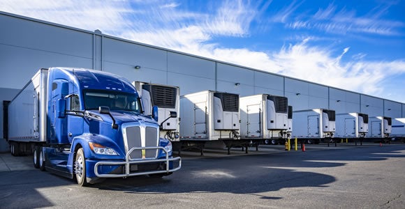 A row of refrigerated trailers, used for specialty shipping, is parked at loading docks outside a large warehouse, with a blue semi-truck in front. The sky above is blue with wispy clouds.