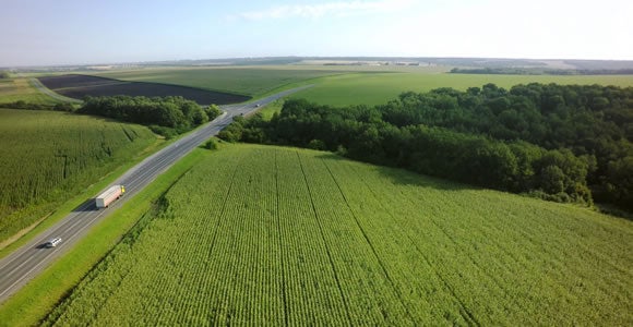 Aerial view of a rural landscape with green fields, a forested area, and a road where a Melon 1 refrigerated freight truck and a car travel under a clear sky.