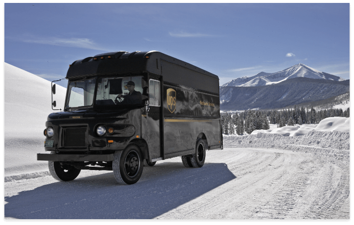 A black UPS delivery truck drives on a snow-covered road with snowy mountains and evergreen trees in the background, reminding us of holiday shipping deadlines under a clear blue sky.