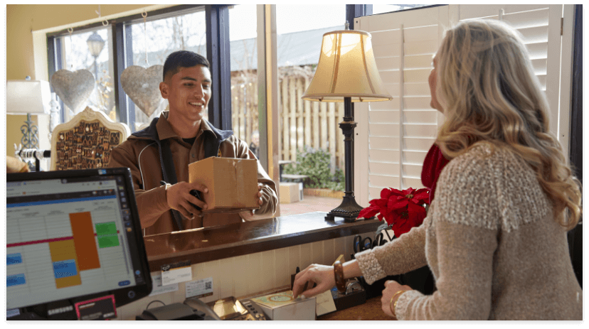 A young delivery person hands a small package to a woman at a reception desk, with a computer monitor and lamp visible—capturing the bright, welcoming atmosphere of holiday shipping as daylight streams through the windows.
